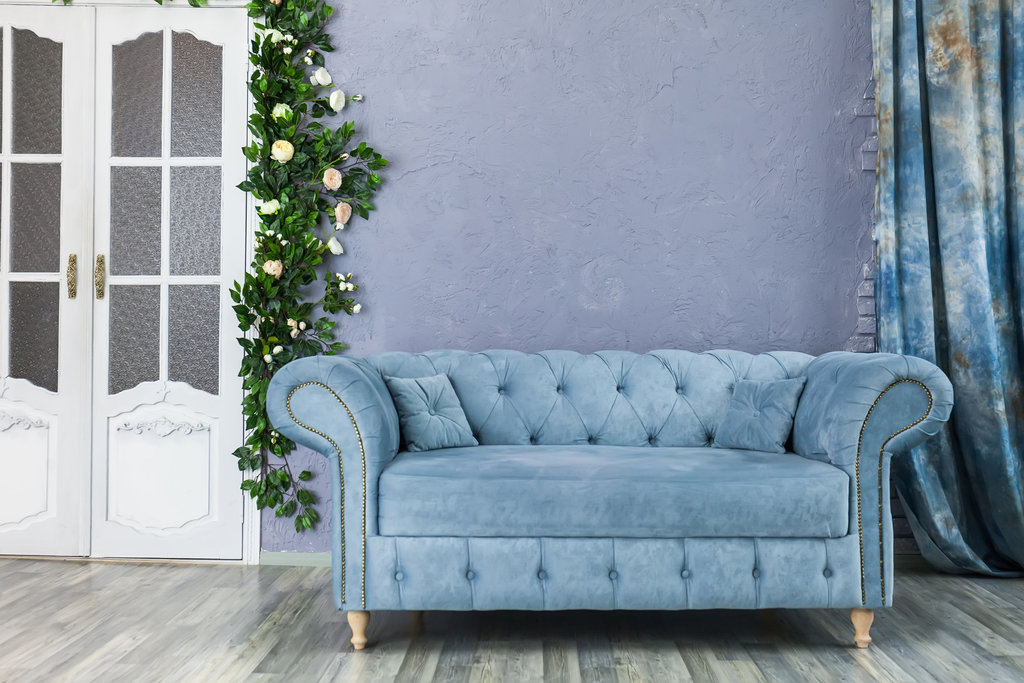 canapé chesterfield bleu pâle posé sur un parquet gris, devant un mur bleu. on y voit sur la gauche une porte blanche à double ouverture ainsi qu'une guirlande de fausses plantes et fleurs retombant sur le côté de la porte.