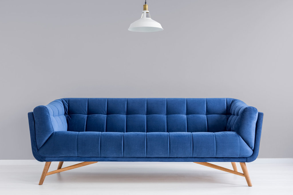 un canapé bleu sur pieds est posé sur un parquet blanc devant un mur lisse gris. un luminaire blanc est suspendu au-dessus du canapé