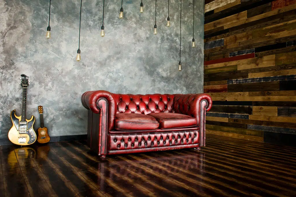 canapé en cuir rouge chesterfield disposé sur un sol et mur en bois. une guitare et un ukulélé sont posés sur le mur gris du fond. des luminaires de style vintage sont également suspendus.
