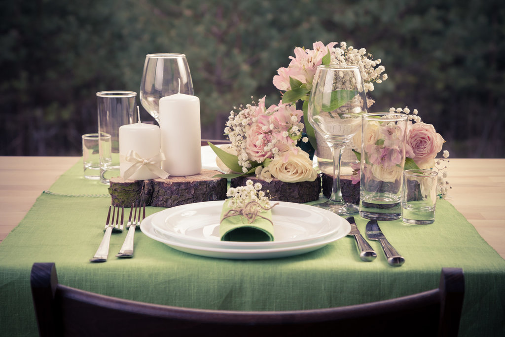 photo d'une décoration de table. sur un chemin de table de couleur verte, sont disposés des couverts, bougies blanches et fleurs rose et blanches