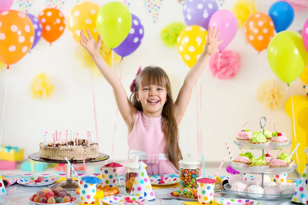 une petite fille en robe rose lève les bras et souris devant sa table d'anniversaire bien décorée de toutes les couleurs. des ballons colorés sont derrière elle