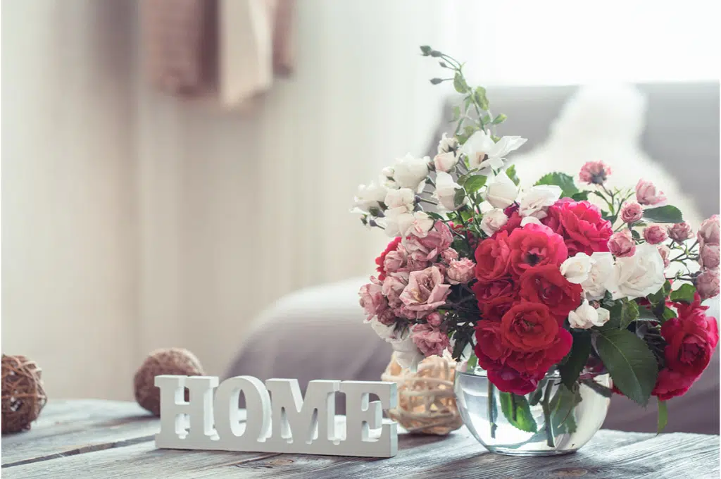 sur une table en bois sont posés un base avec des fleurs rouges, roses et blanches ainsi que un élément en bois blanc écrit "Home".