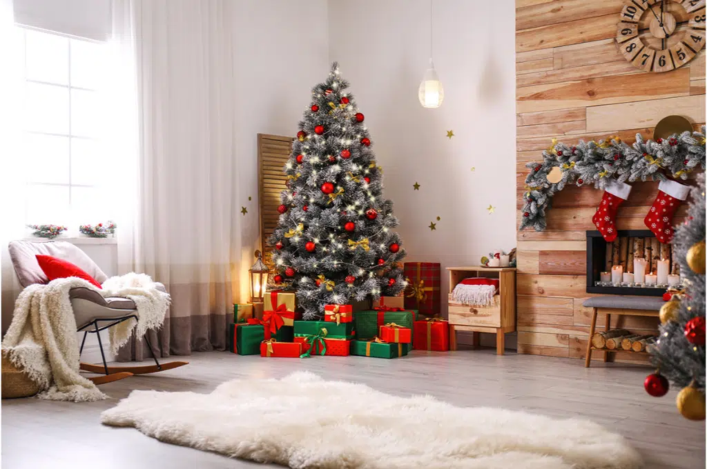 dans un salon décoré pour Noël avec un sapin et des cadeaux au pied du sapin. un tapis en peau d'animal blanc est posé sur le sol en parquet gris. un fauteuil qui se balance a un plaid blanc douillet posé dessus ainsi qu'un coussin rouge. des chaussettes de Noël sont accrochées au-dessus de la cheminée.