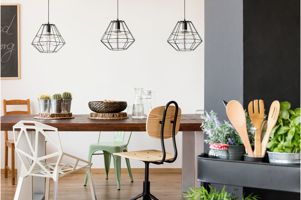 dans une cuisine, des lampes métalliques sont suspendues au-dessus d'une table en bois avec des chaises de styles très différents. on voit sur le côté, divers ustensiles de cuisine ainsi que des plantes aromatiques.