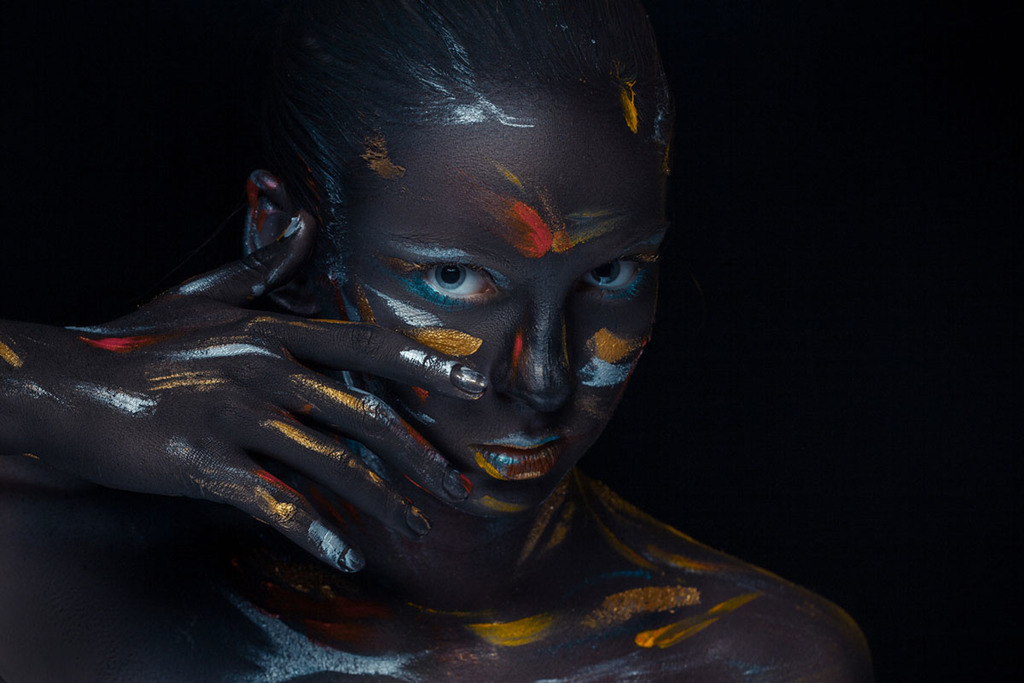 sur fond noir et dans une ambiance sombre, une femme prend la pose avec de la peinture de couleurs sur le visage, la main et ses épaules.