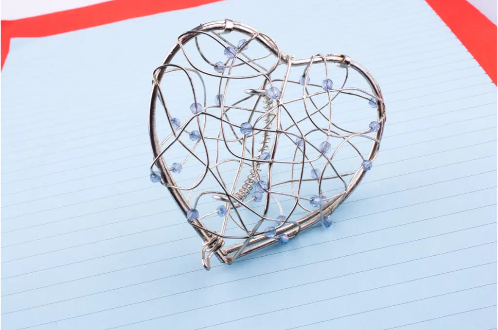 sur une feuille de papier avec des traits, est posé un coeur en fil de fil. de nomreuses petites perles bleues sont de part et d'autres enfilées dans les fils de fer.