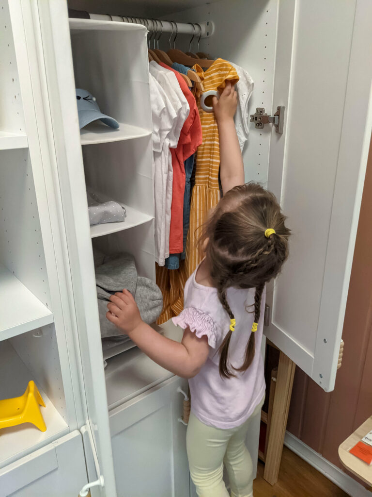 une jeune fille touche ses vêtements dans son armoire à vêtements qui est ouverte. on n'y voit notamment des robes sur cintres.