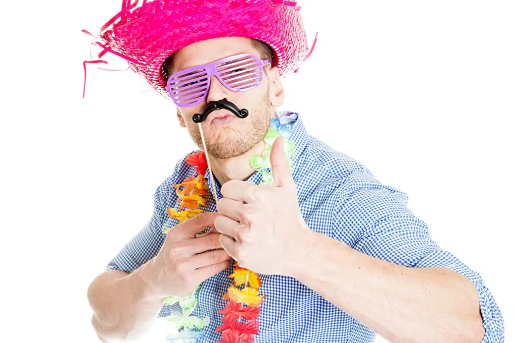 un homme déguisé avec un chapeau de paille rose, un collier de fleurs tahitien, des lunettes en plastique violettes, tient une moustache noire en plastique. son autre main fait un pouce en l'air.