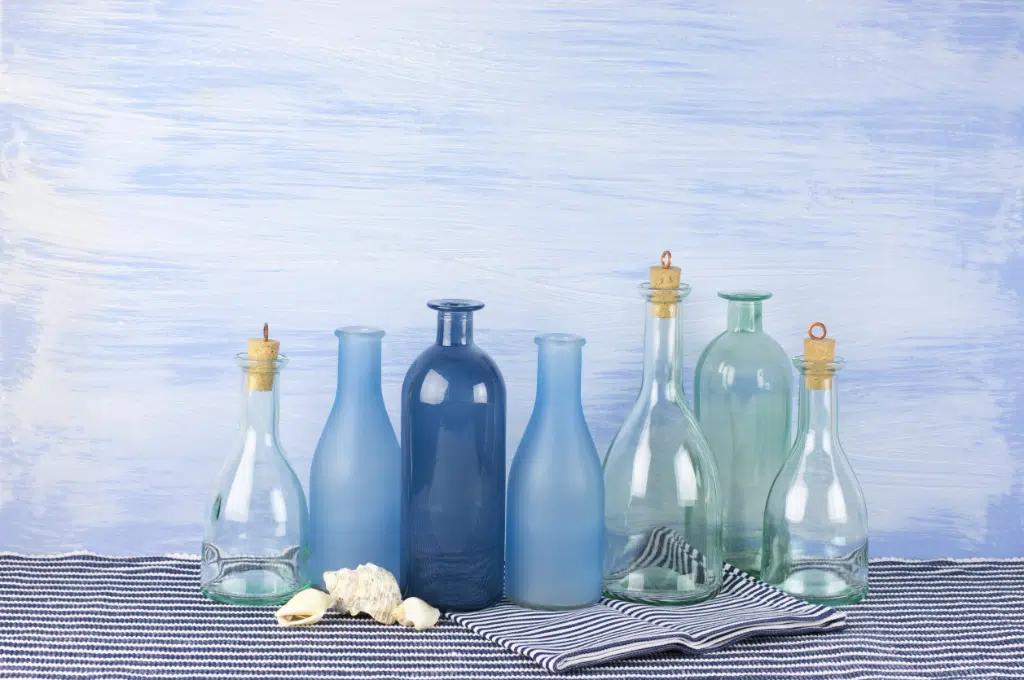 de jolies bouteilles en verre colorées ou transparentes sont alignées devant un mur et posé sur un tapis rayé bleu et blanc. Des coquillages sont posés devant.