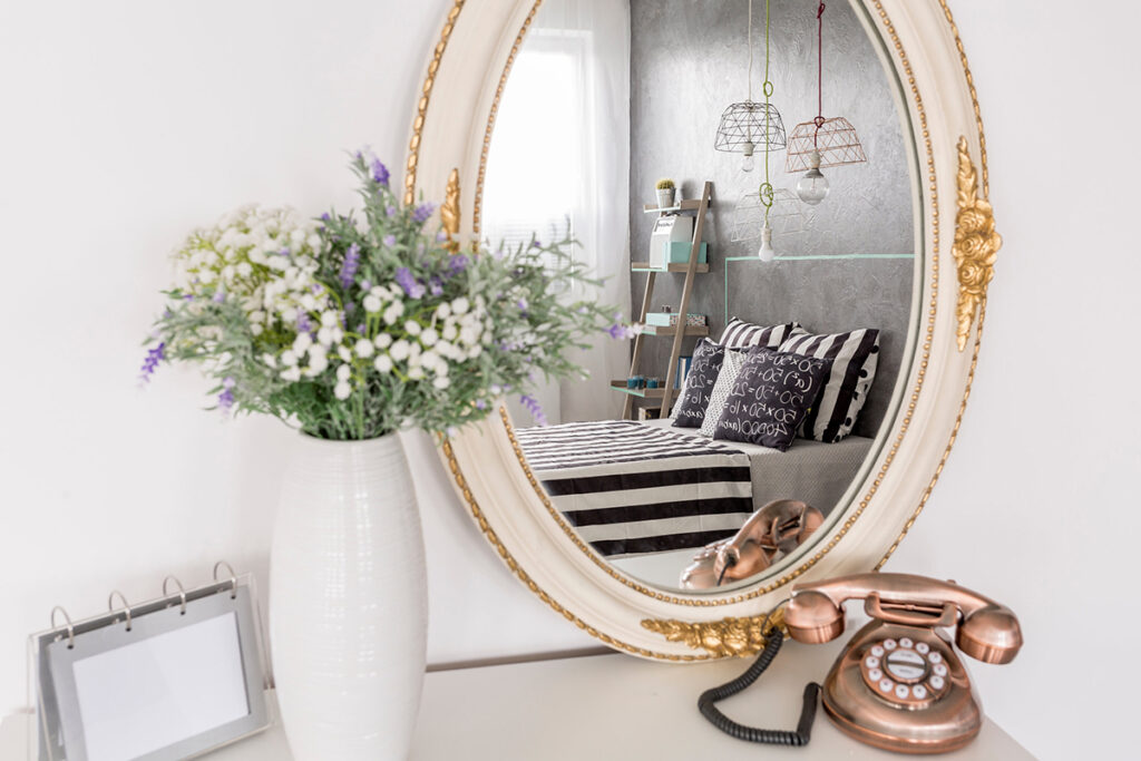 un miroir ovale est dans une chambre posé sur un meuble. un téléphone rétro est à côté ainsi qu'un vase blanc avec des fleurs