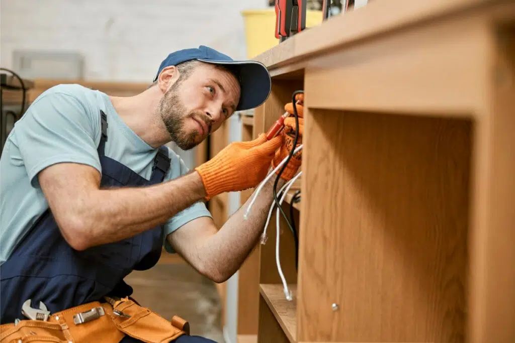 un électricien en salopette bleue et casquette bleue installe des fils électriques à l'arrière d'un bureau en bois. il porte des gant orange et utilise un outils sur des fils élctriques.