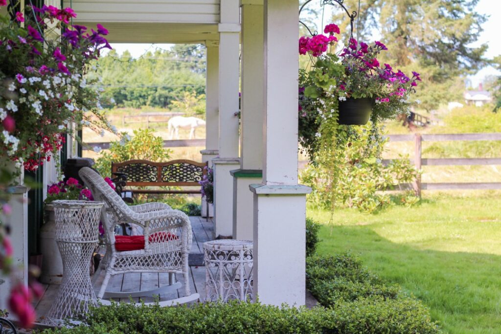 à l'entrée d'une maison, divers pots de fleurs sont suspendus. on distingue un siège à bascule en rotin black ainsi qu'un ensemble de tables d'appoint de la même gamme.