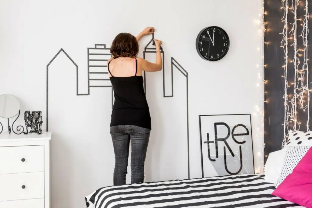 une femme en débardeur noir et en jean, de dos, pose un stickers sur un mur blanc. le stickers a la forme de buildings. une horloge est également accrochée au mur.