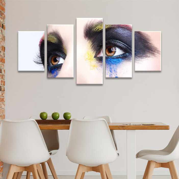 Tableau visage au regard maquillé accroché au mu clair d'une cuisine avec une longue table en bois sur laquelle se trouvent trois pommes vertes et entourée de chaises aux dossiers blancs