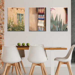 Tableau Mexique vie quotidienne accroché au mur blanc d'une cuisine avec mur en pierre et table en bois clair entourée de chaises blanches au design épuré.